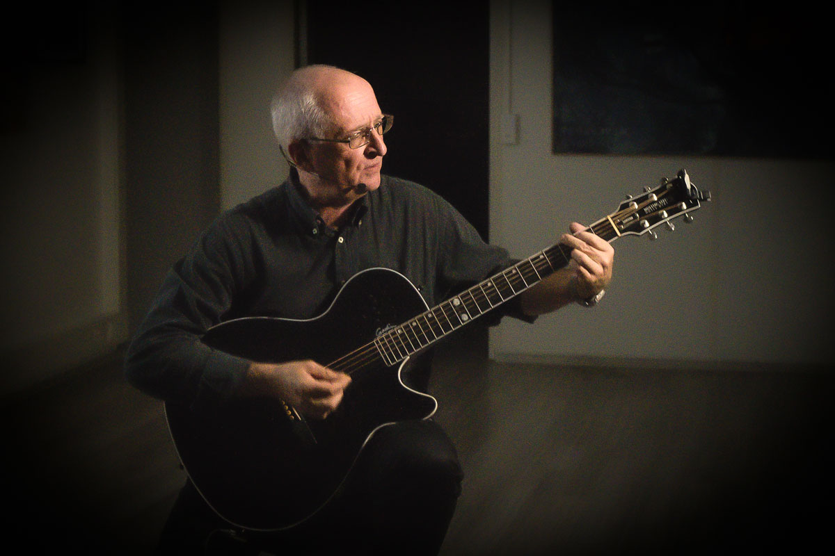 Enrico Pestalozzi en concert, assis, jouant de la guitare acoustique et chantant au micro, format voix/guitare